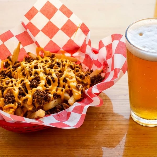 Chili cheese fries in a red basket with checkered paper, next to a glass of beer.