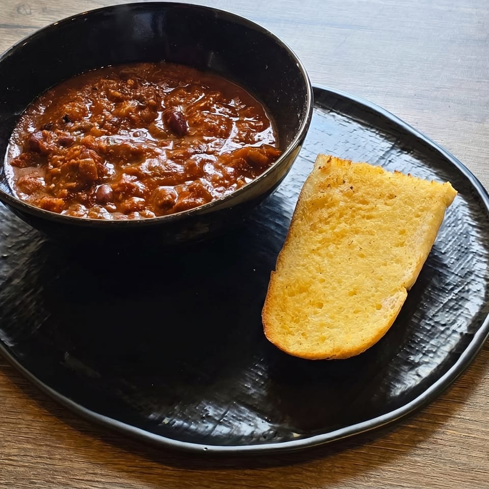 A black bowl of chili with a slice of golden cornbread on a black plate.