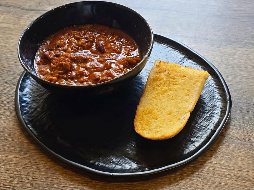 A black bowl of chili with a slice of golden cornbread on a black plate.