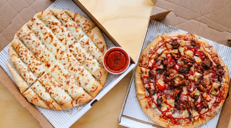 Cheesy breadsticks with marinara sauce and a BBQ chicken pizza with pineapple in takeout boxes.